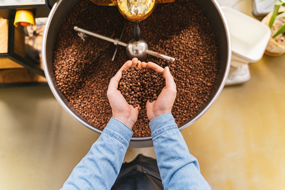 A person holds fresh coffee beans over a large roasting bowl in a coffee shop